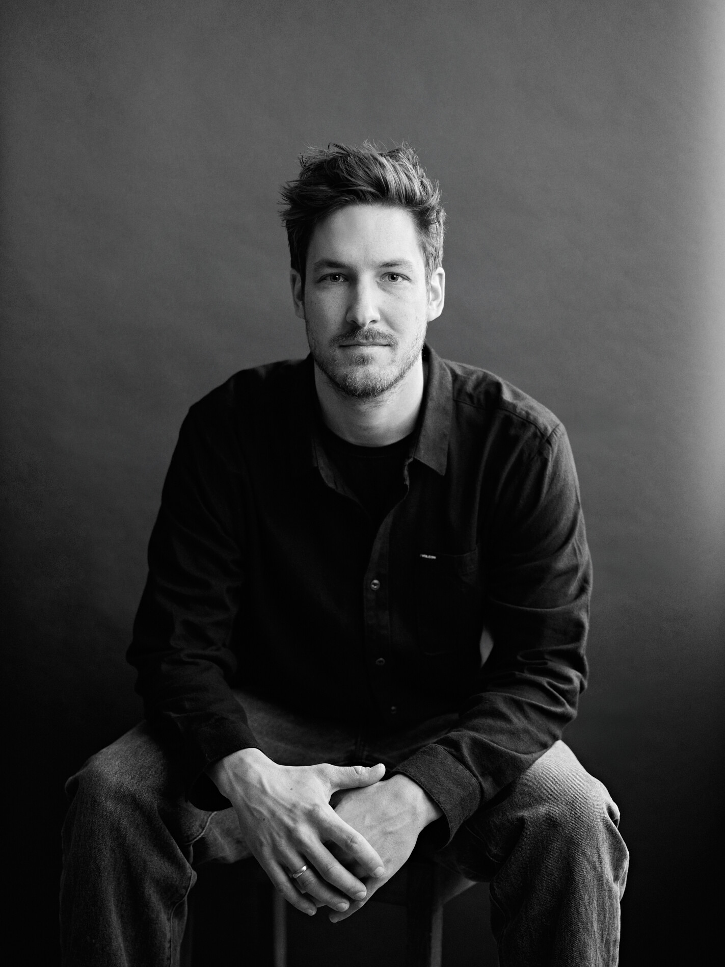 Eine Person sitzt auf einem Hocker vor einem dunklen Hintergrund, trägt ein dunkles Hemd und Jeans und blickt mit einer Intensität in die Kamera, die die Essenz der Porträtfotografie einfängt. Dieser fesselnde Moment wurde von einem talentierten Fotografen in Düsseldorf gekonnt festgehalten. Portrait und Reportage Fotografie für Corporate und Editorial