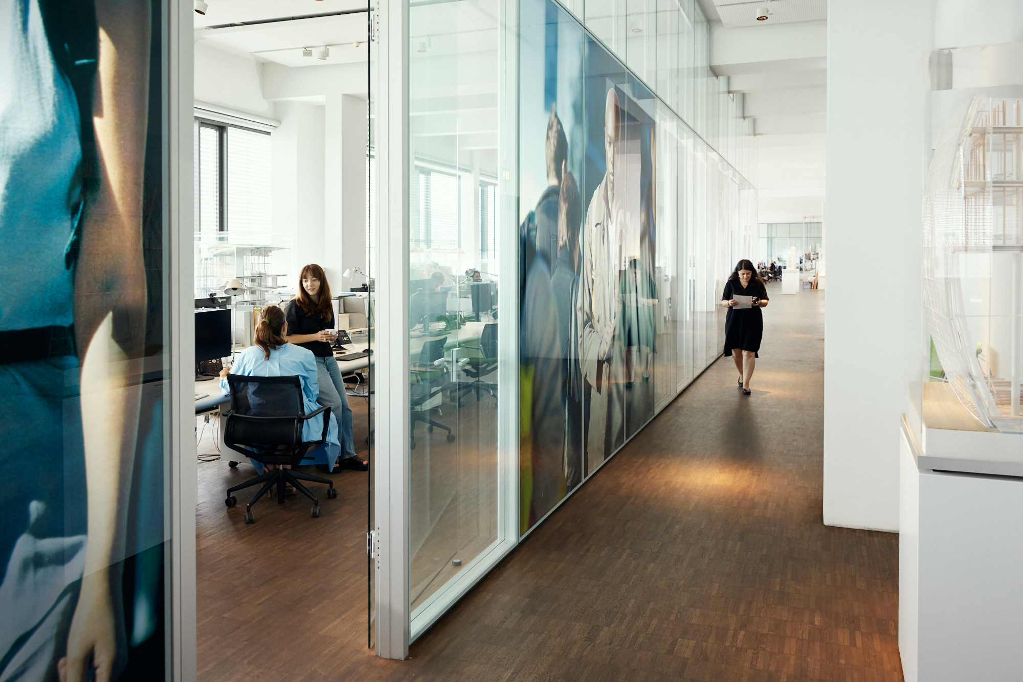 Ein moderner Büroflur mit Glaswänden und großen Personenbildern bietet eine beeindruckende Kulisse. In diesem Büro sitzen zwei Frauen konzentriert an ihren Schreibtischen, während eine andere durch den Flur schreitet und die Essenz der Unternehmensfotografie verkörpert. Portrait und Reportage Fotografie für Corporate und Editorial