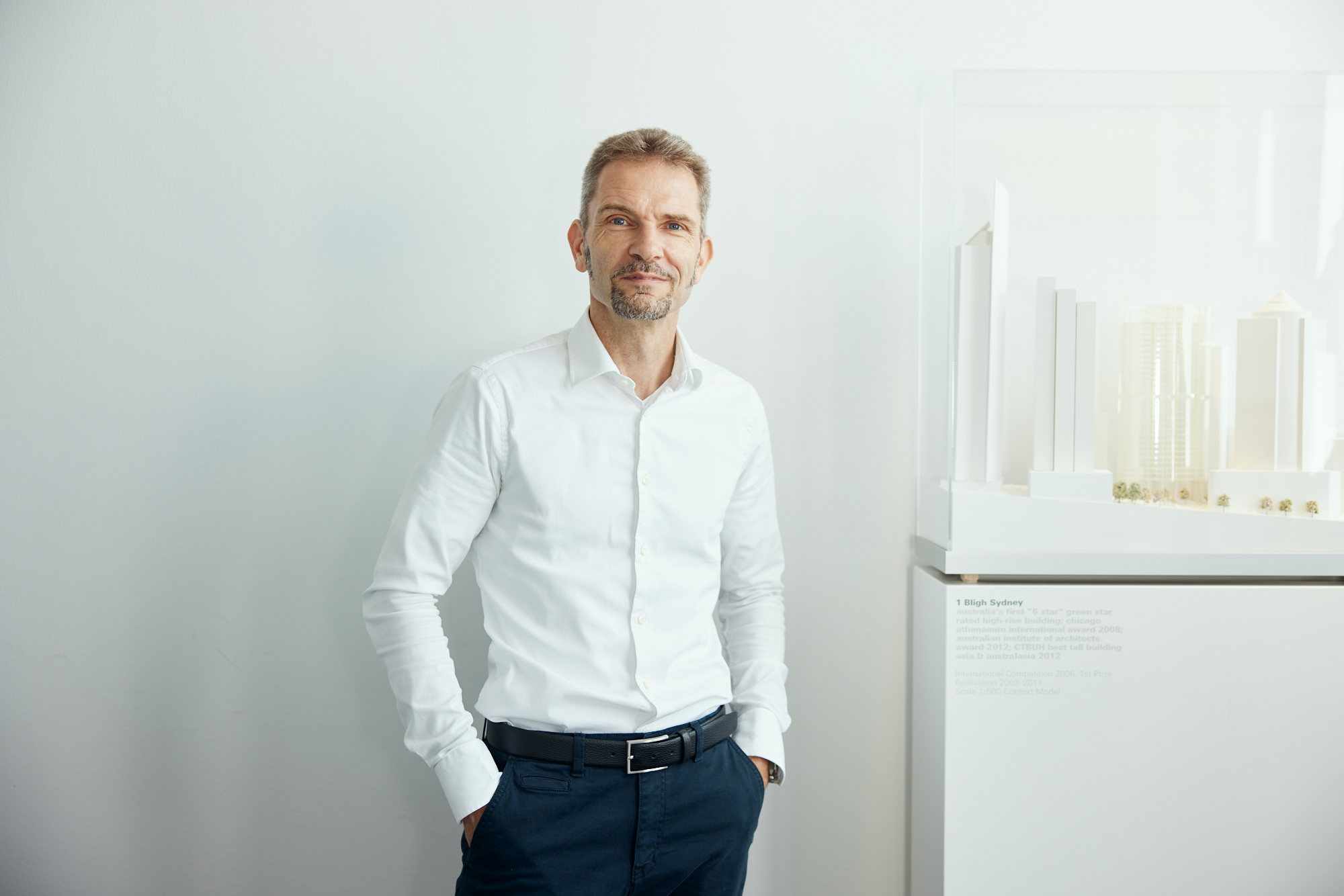 Porträt eines Mannes in weißem Hemd und dunkler Hose, der neben einem Architekturmodell in einem hellen Raum steht und die Essenz der Unternehmensfotografie einfängt. Portrait und Reportage Fotografie für Corporate und Editorial