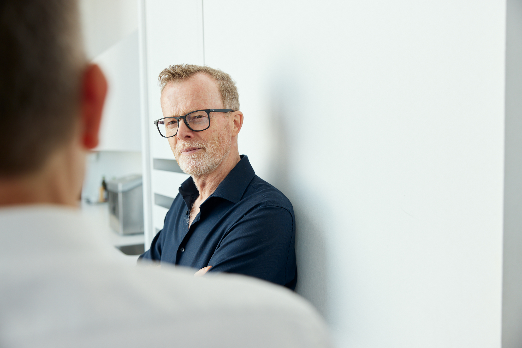 Ein Mann mit Brille und dunklem Hemd steht selbstbewusst mit verschränkten Armen da, was an ein Firmenporträt erinnert, während er im Vordergrund einer hellen Innenumgebung mit einer anderen Person interagiert. Portrait und Reportage Fotografie für Corporate und Editorial