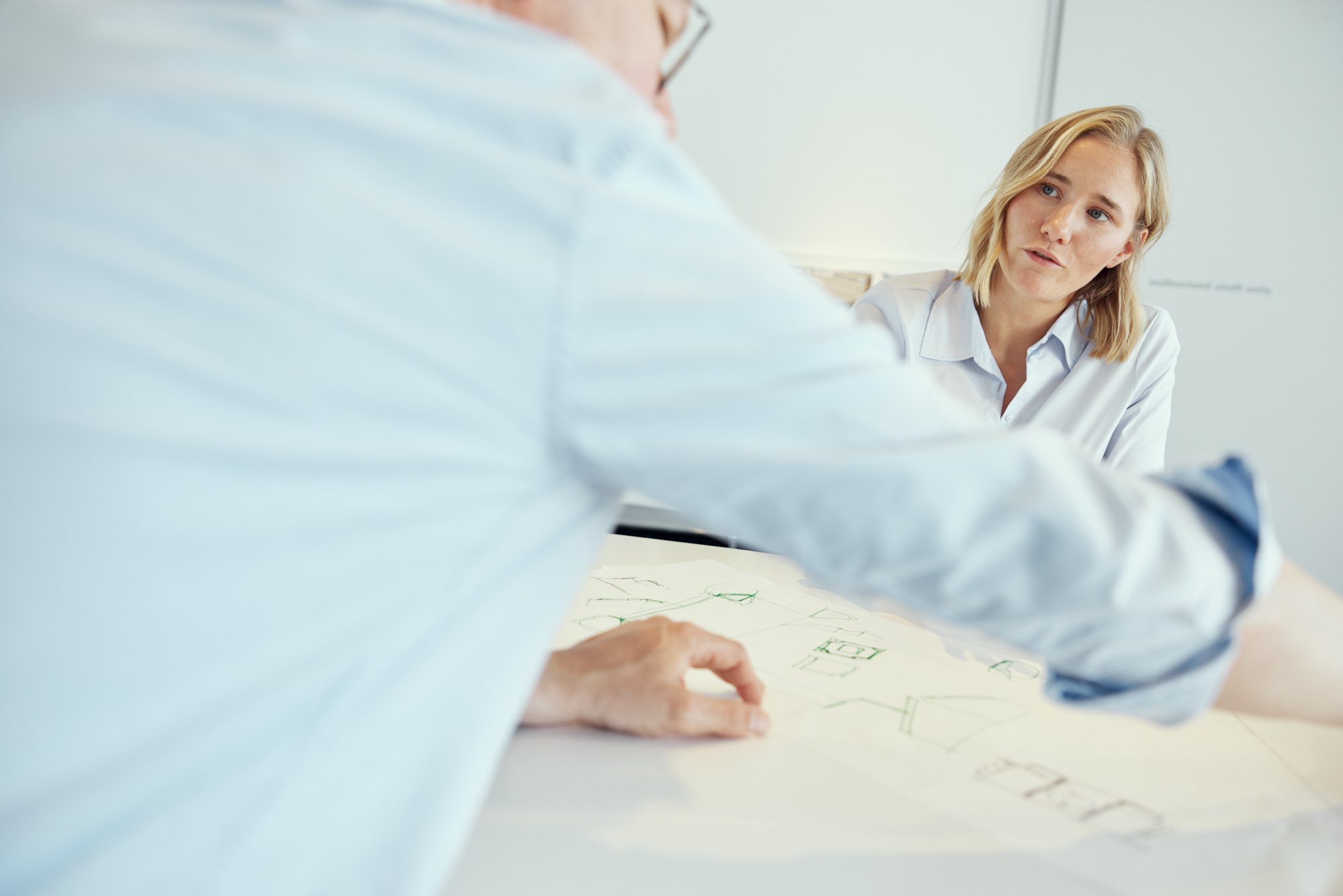 In einer dynamischen Präsentation von Unternehmensfotografie sitzen zwei Kollegen an einem Tisch mit Architekturplänen. Die Frau achtet aufmerksam darauf, während der Mann auf Details hinweist – ein Beispiel für Teamarbeit in Düsseldorf. Portrait und Reportage Fotografie für Corporate und Editorial