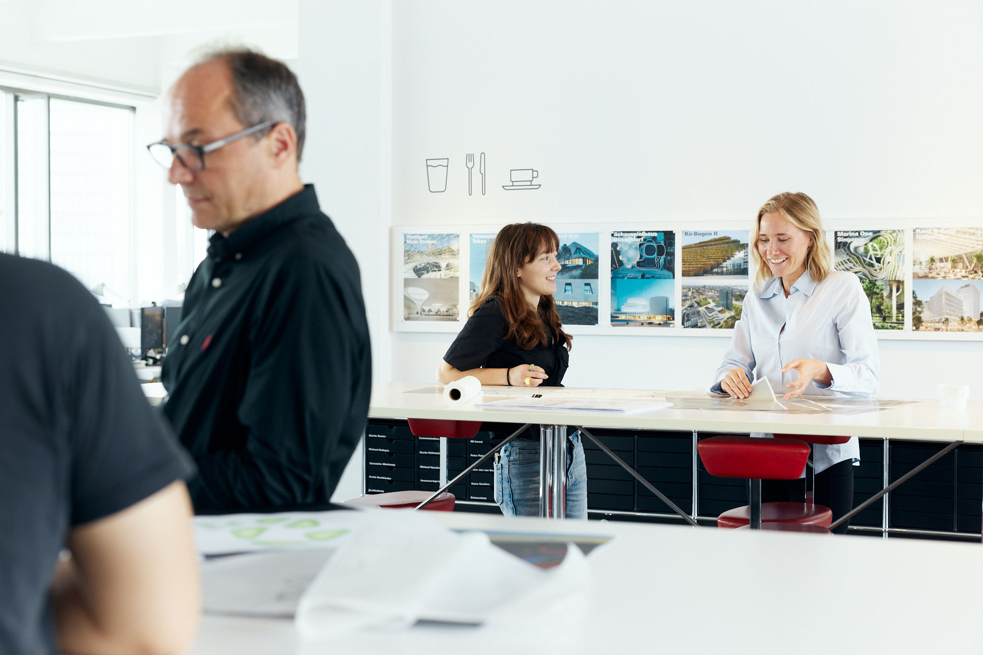 In einem Düsseldorfer Büro werden drei Personen für einen Moment der Unternehmensfotografie festgehalten; zwei sitzen lächelnd und unterhalten sich mit Papieren an einem Tisch, während eine dritte im Vordergrund steht und Dokumente durchsieht. Portrait und Reportage Fotografie für Corporate und Editorial