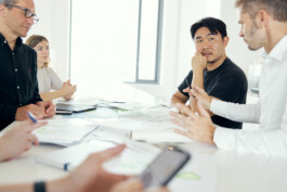 Im Besprechungsraum diskutiert eine bunt gemischte Gruppe an einem mit Papieren übersäten Tisch. Während einige Notizen machen, fotografiert einer mit seinem Smartphone und fängt so die Essenz der Unternehmensfotografie perfekt ein. Es ist eine dynamische Szene mitten im Herzen Düsseldorfs. Portrait und Reportage Fotografie für Corporate und Editorial