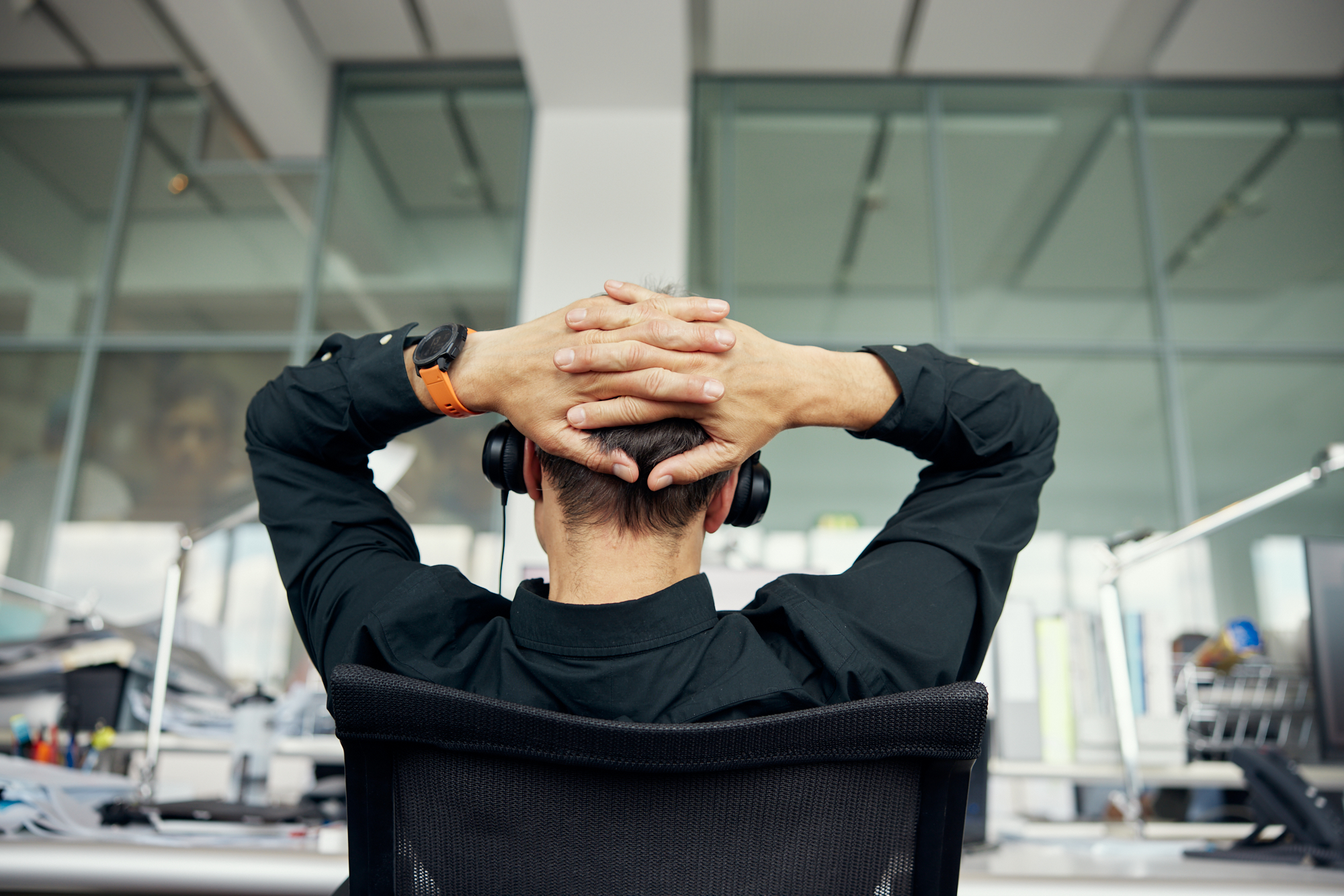 In einem Firmenbüro sitzt eine Person im schwarzen Hemd auf einem Bürostuhl, die Hände hinter dem Kopf verschränkt und Kopfhörer auf. Die Glaswand und die Schreibtische mit Büroausstattung vermitteln die elegante Atmosphäre von Unternehmensfotografie mitten in Düsseldorf. Portrait und Reportage Fotografie für Corporate und Editorial