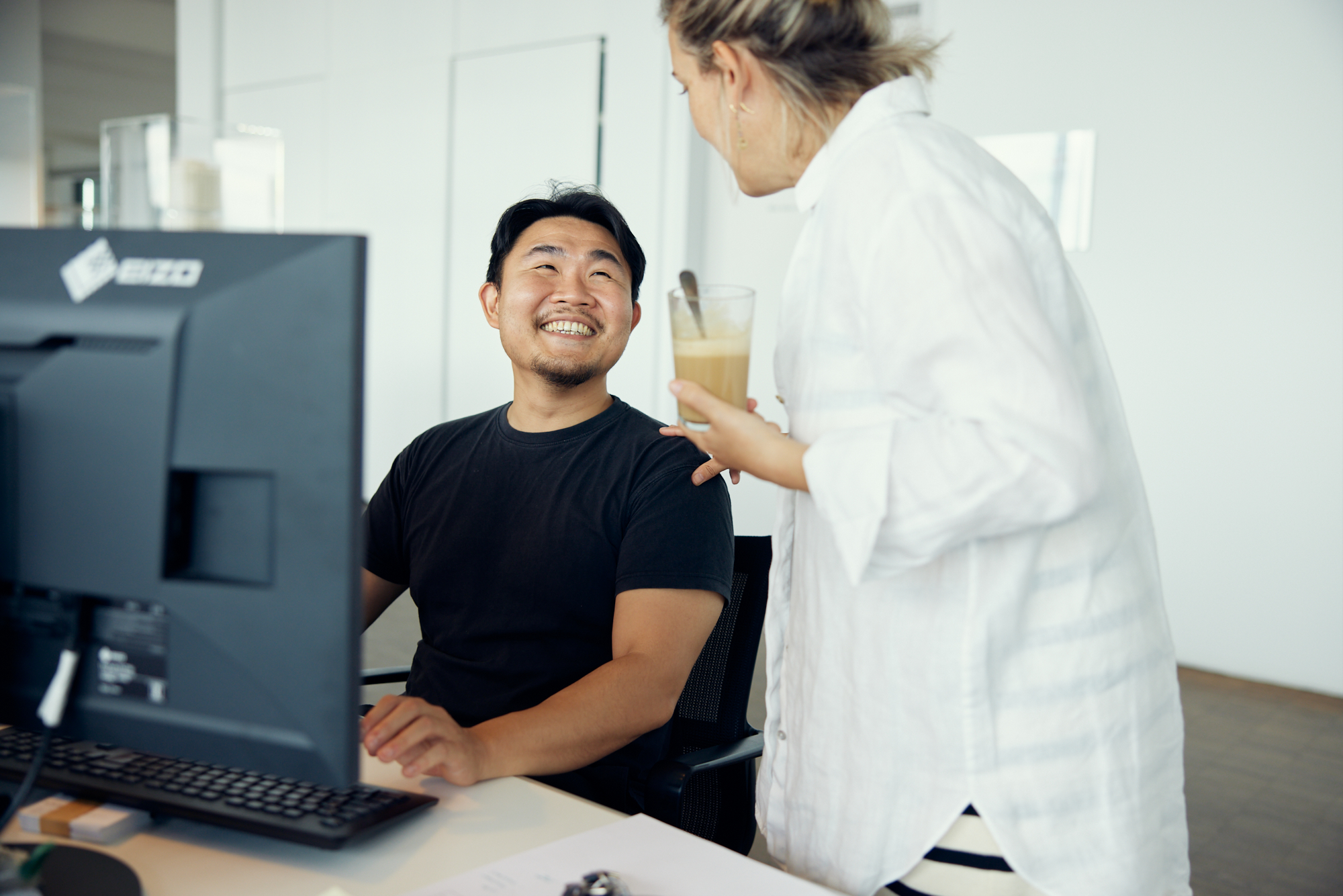 In einem Düsseldorfer Büro sitzt ein Mann an einem Schreibtisch mit einem Computer und lächelt eine Frau mit einem Drink an. Sie unterhalten sich angeregt und fangen so die Essenz der Unternehmensfotografie ein und heben gleichzeitig echte Verbindungen hervor. Portrait und Reportage Fotografie für Corporate und Editorial