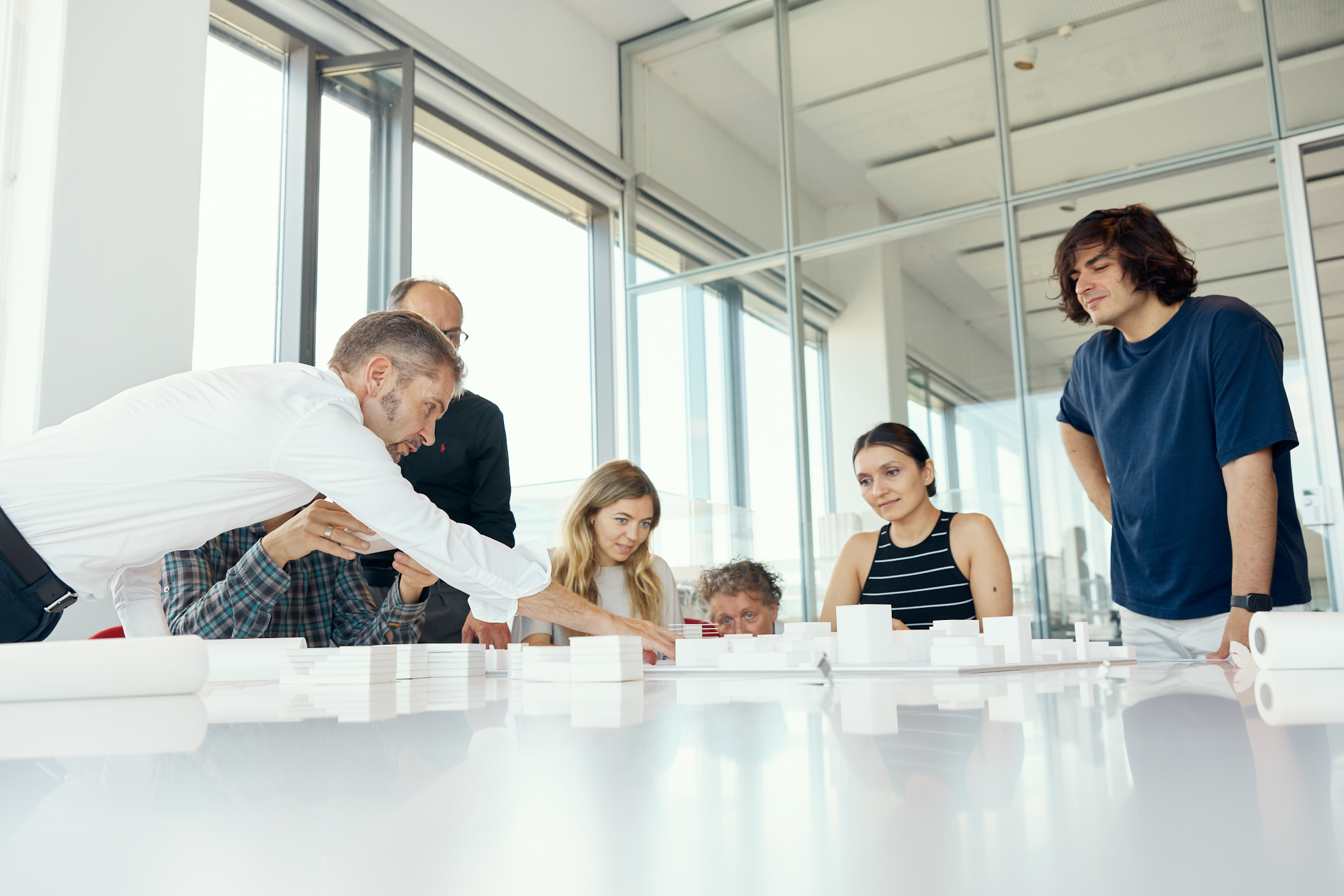 In einem hellen Düsseldorfer Büro versammelt sich eine bunt gemischte Gruppe um einen Tisch, diskutiert und arbeitet enthusiastisch an einem weißen Architekturmodell und fängt so die Essenz der Unternehmensfotografie ein. Portrait und Reportage Fotografie für Corporate und Editorial