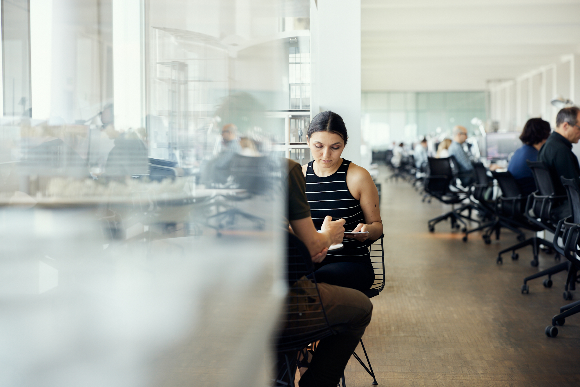 Eine Frau im gestreiften Oberteil sitzt mit einer weiteren Person an einem Tisch in einem hellen, offenen Büro in Düsseldorf. Im Hintergrund arbeiten mehrere Personen an Schreibtischen und fangen mit authentischen Mitarbeiterporträts die Essenz der Unternehmensfotografie ein. Portrait und Reportage Fotografie für Corporate und Editorial