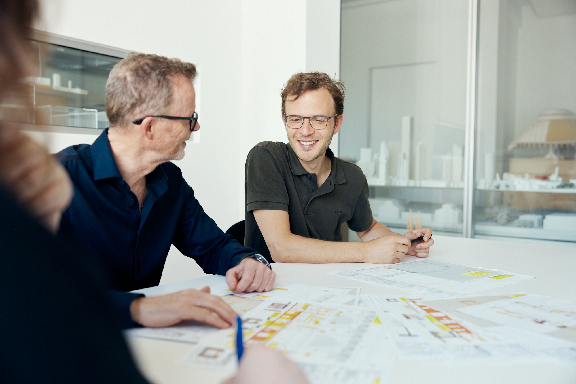 Drei Personen sitzen an einem Tisch in Düsseldorf und diskutieren Architekturpläne. Zwei prominent abgebildete Männer unterhalten sich lächelnd. Vor ihnen liegen Pläne und Skizzen ausgebreitet, die durch Unternehmensfotografie die Essenz professioneller Zusammenarbeit einfangen. Portrait und Reportage Fotografie für Corporate und Editorial