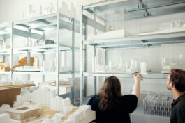 Zwei Personen betrachten Architekturmodelle auf eleganten Metallregalen und fangen so die Essenz des Bürodesigns in einem Raum voller Miniatur-Stadtansichten und Gebäudeentwürfe ein, wodurch ein professioneller Eindruck entsteht, der an hochwertige Unternehmensfotografie erinnert. Portrait und Reportage Fotografie für Corporate und Editorial